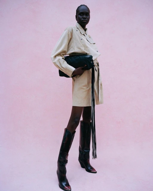 Full-length portrait of an adult female fashion model posing in a studio with a clutch purse, wearing a belted dress and black western boots.