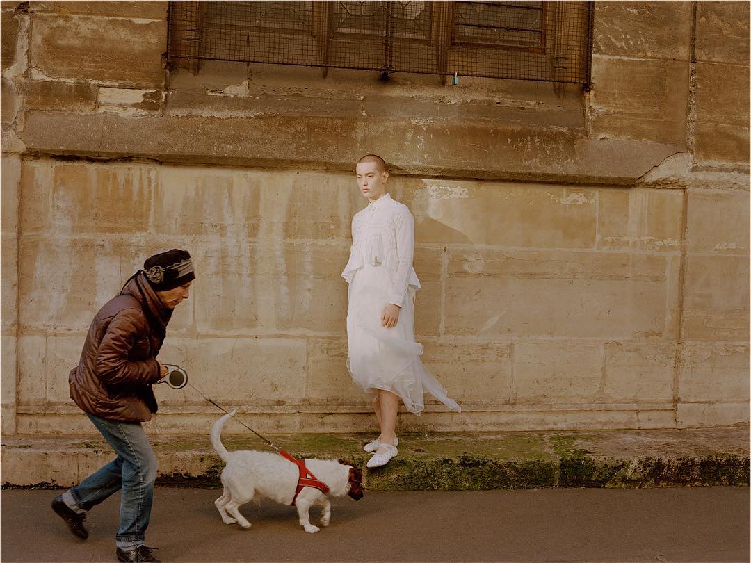 Woman in white dress standing near stone wall, another person walking dog on leash, urban setting, neutral colors, diffused light.