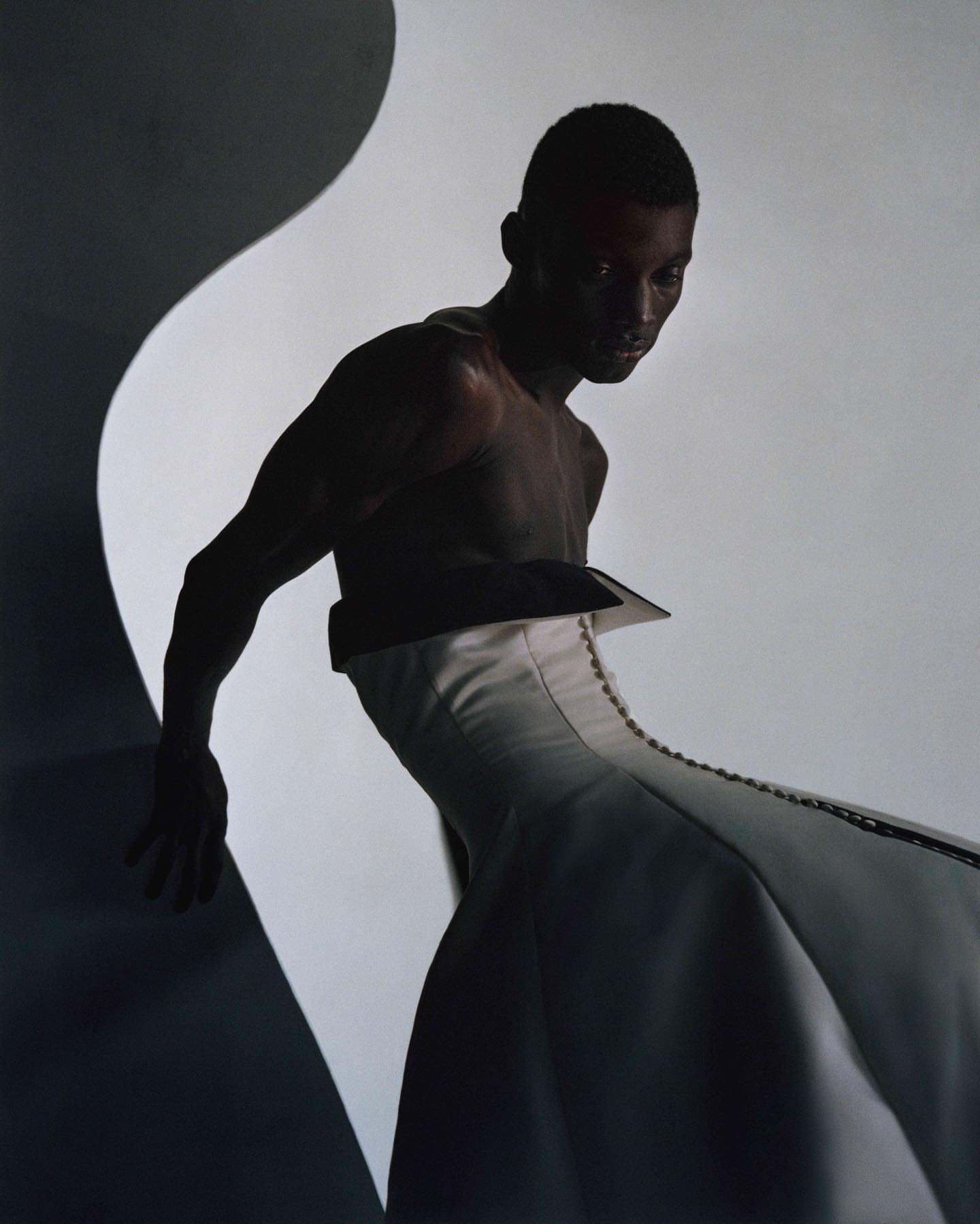 A shirtless Black male model in a white, structured garment poses in a studio with diffused lighting and a dark, curved background element.
