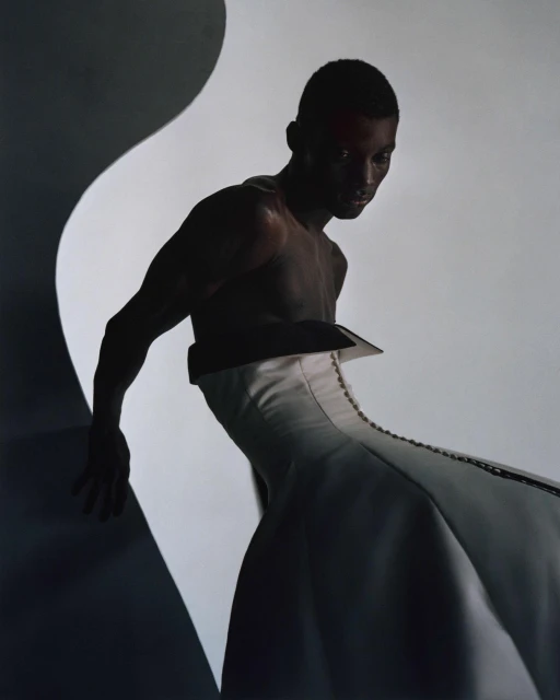 A shirtless Black male model in a white, structured garment poses in a studio with diffused lighting and a dark, curved background element.