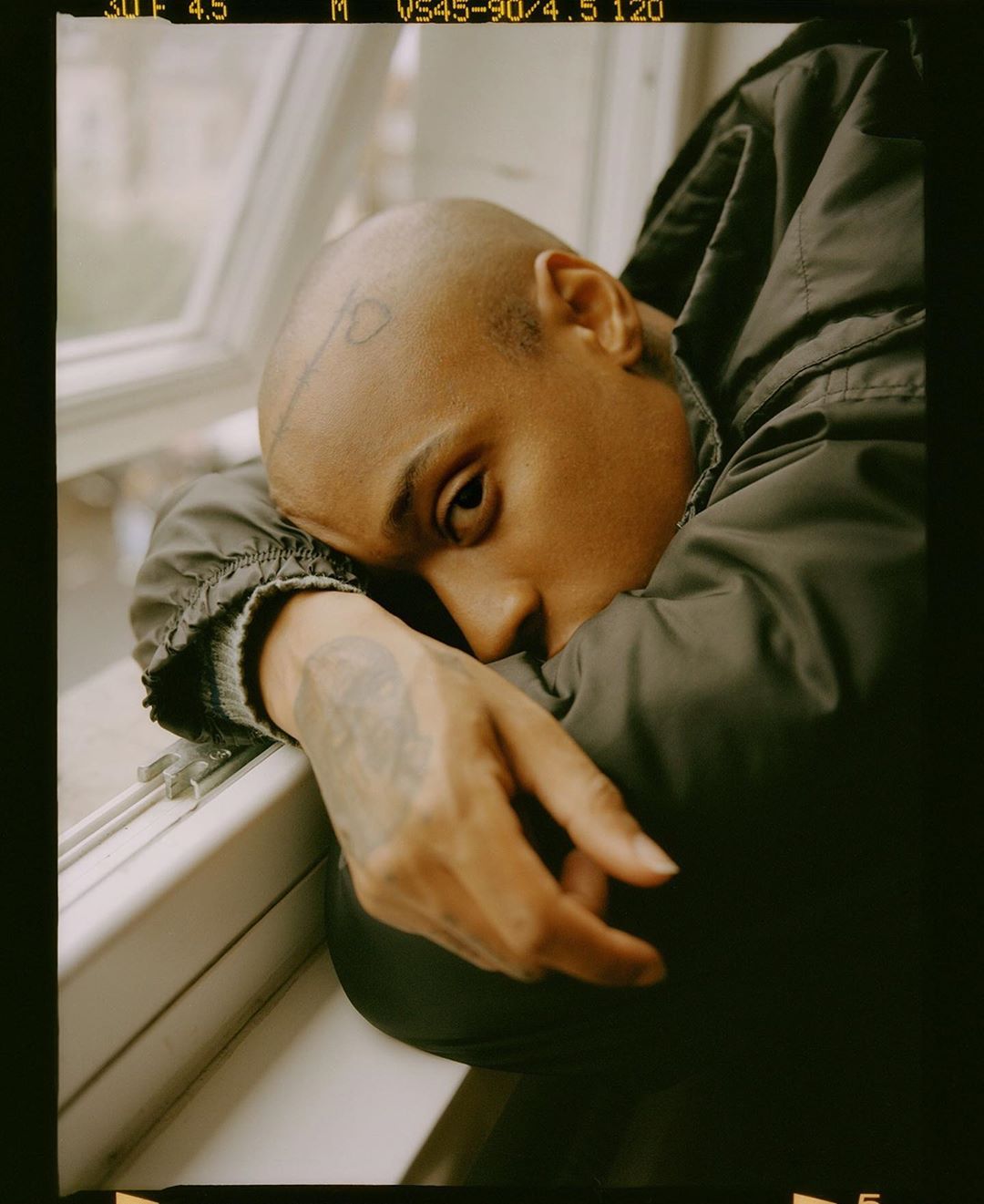 Close-up portrait of a bald adult leaning against a window frame, wearing a dark jacket in an indoor setting with natural lighting.