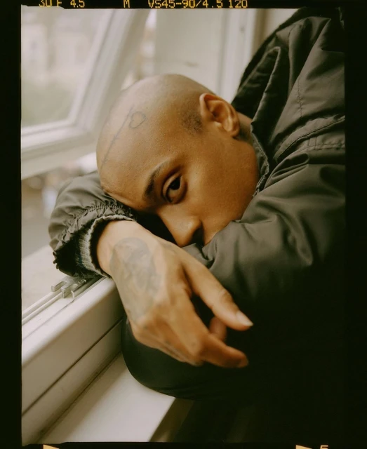 Close-up portrait of a bald adult leaning against a window frame, wearing a dark jacket in an indoor setting with natural lighting.