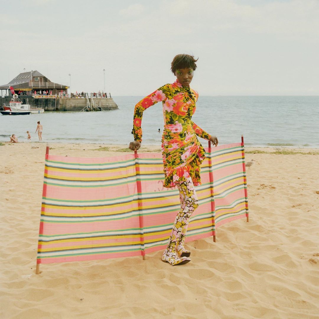 A Black female model wearing a floral dress and leggings poses on a beach next to a striped windbreak, with a pier and the sea in the background.