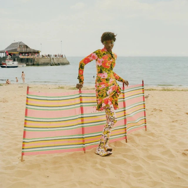 A Black female model wearing a floral dress and leggings poses on a beach next to a striped windbreak, with a pier and the sea in the background.