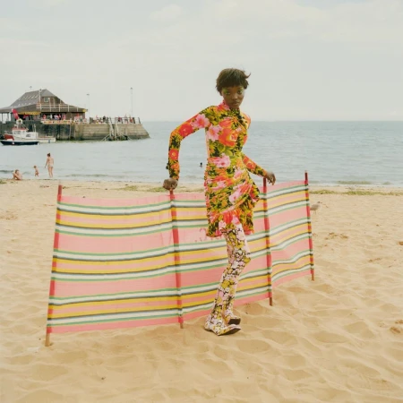 A Black female model wearing a floral dress and leggings poses on a beach next to a striped windbreak, with a pier and the sea in the background.