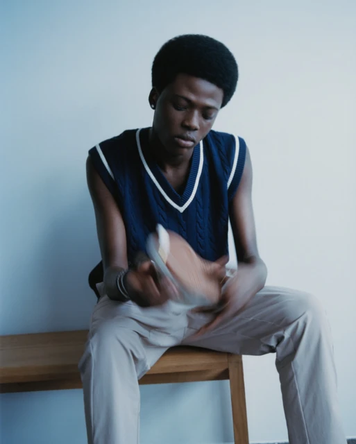 A young Black adult sits on a wooden bench indoors, holding a beige shoe in his hands, wearing a navy blue sweater vest and khaki trousers.