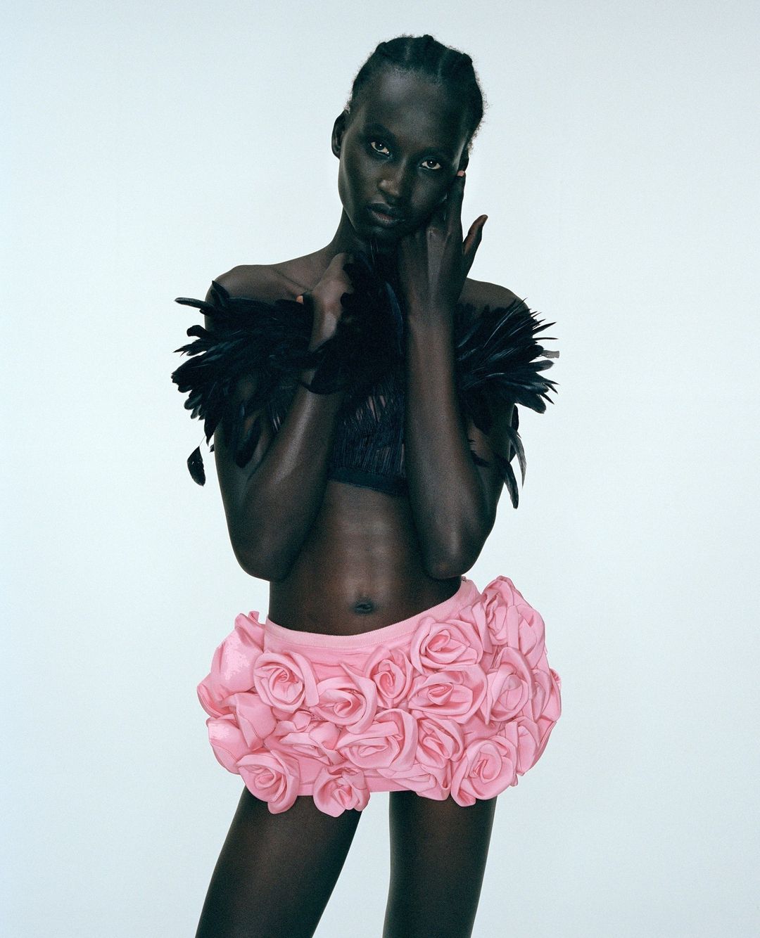 Fashion shot of a dark-skinned model wearing a feathered top and a pink rose-covered skirt posing against a neutral background.