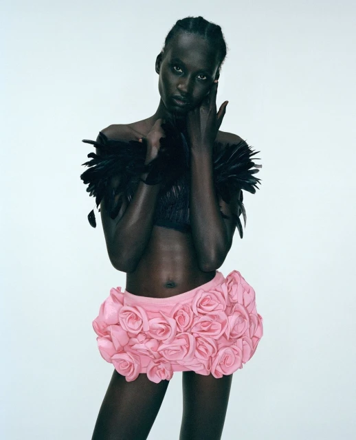 Fashion shot of a dark-skinned model wearing a feathered top and a pink rose-covered skirt posing against a neutral background.