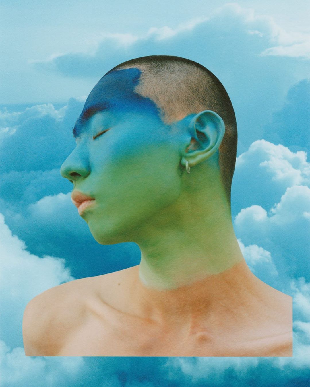 Portrait of a person with blue and green face paint against a cloudy sky background. Eyes are closed in a serene profile.