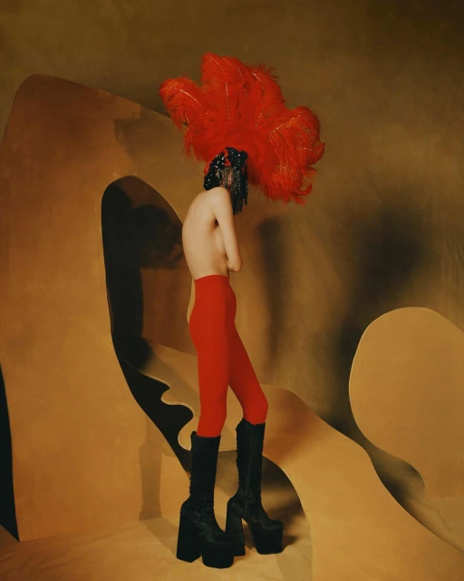 Artistic fashion photo of a person in red tights and platform boots with a large red feather headpiece against a tan backdrop.