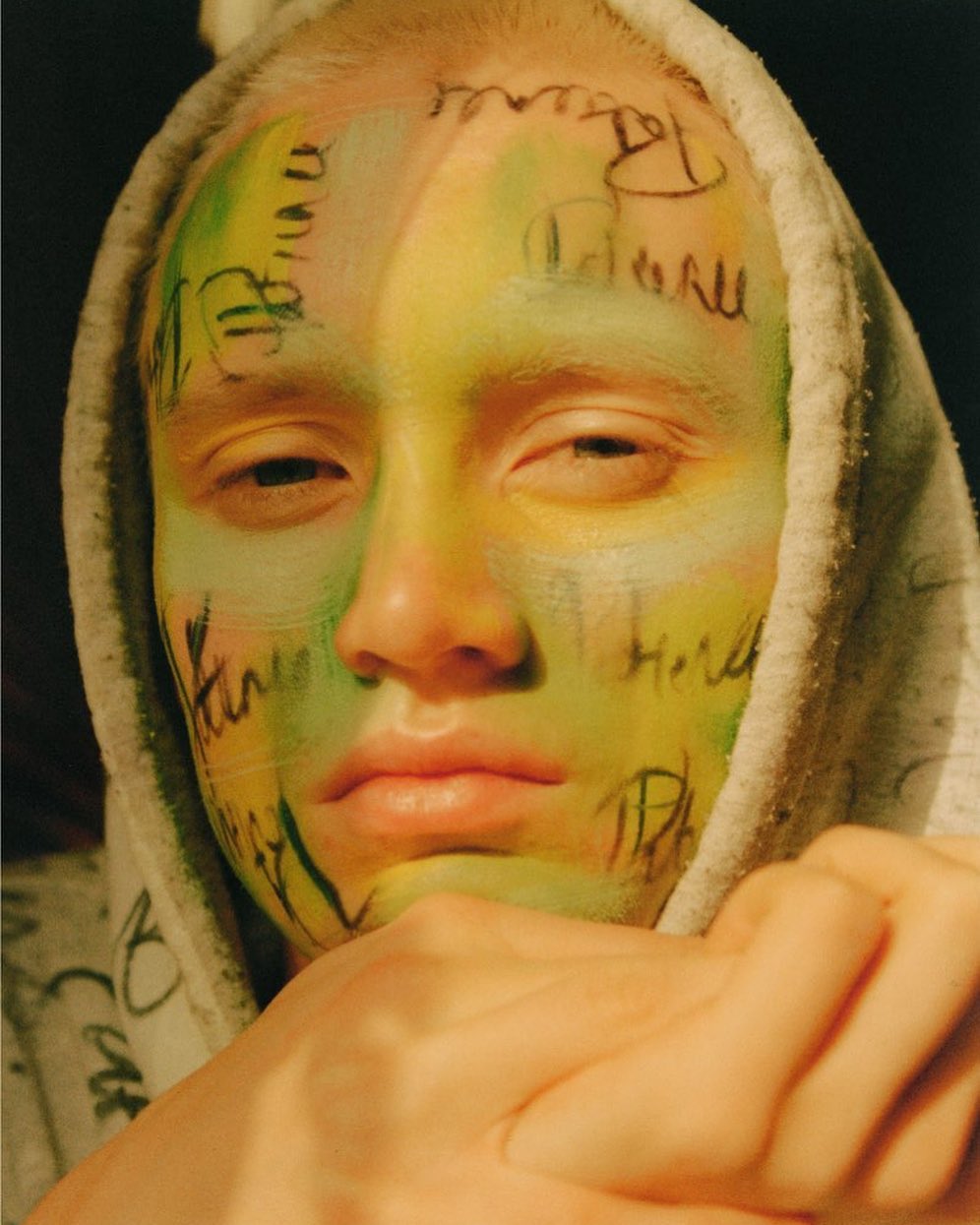 Close-up portrait of a young adult with unique face paint and a hooded sweatshirt, set against a dark background in a photo studio.