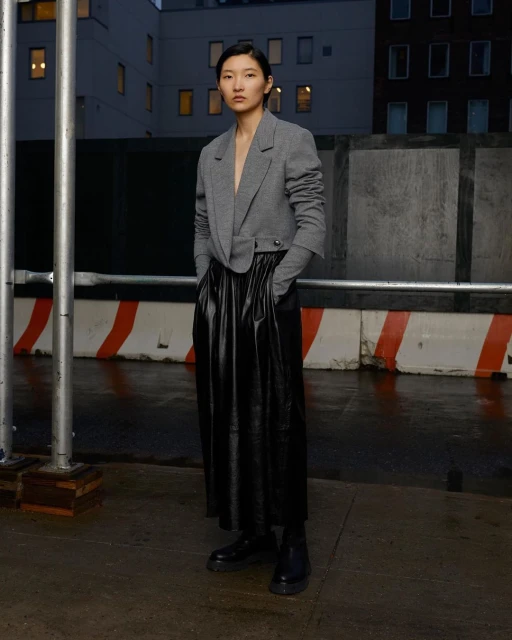 Asian woman in a gray blazer and long black skirt poses against an urban backdrop in an editorial fashion shot at dusk.