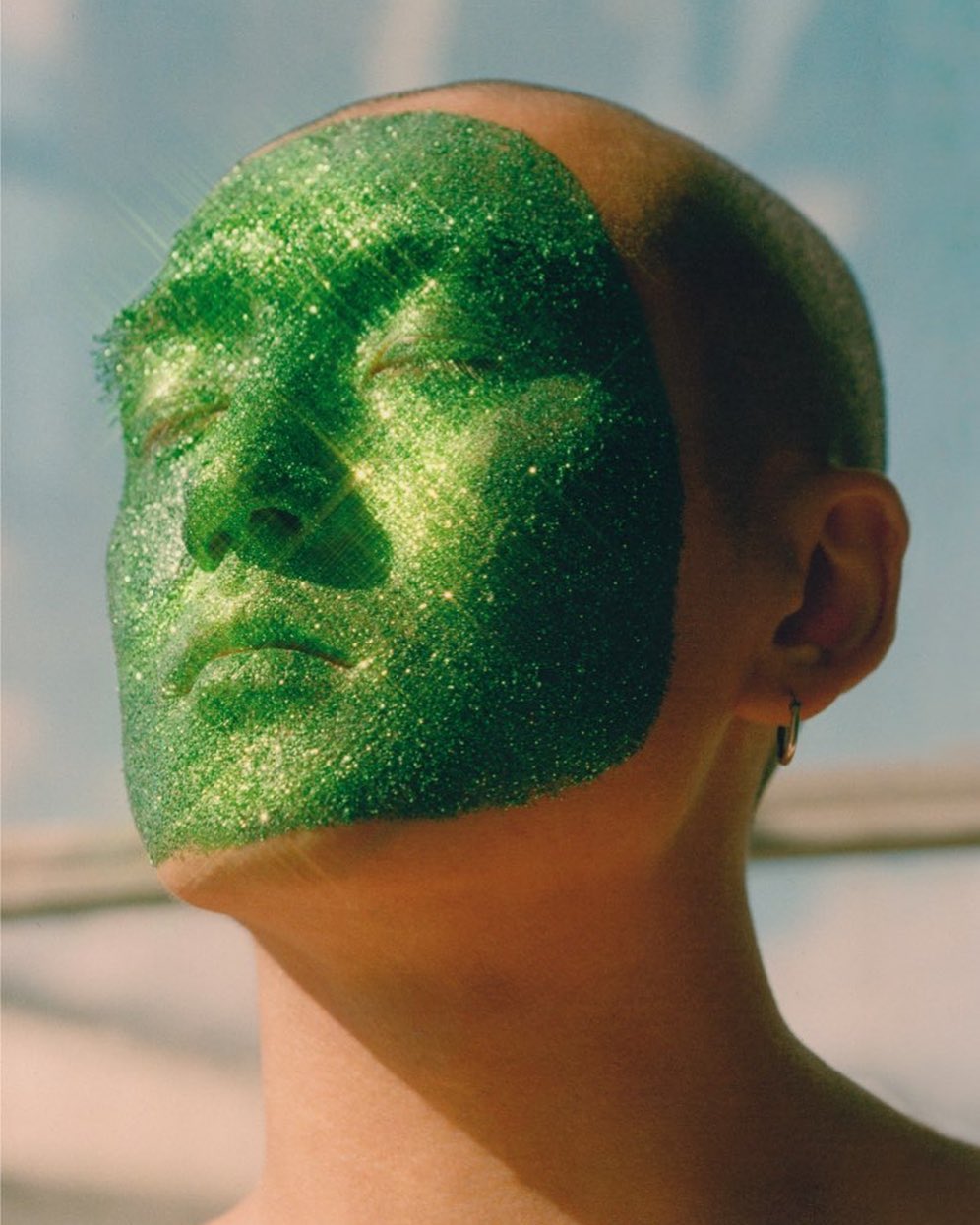 Close-up portrait of a bald person with closed eyes and green glitter covering their face, against a blurred blue sky.