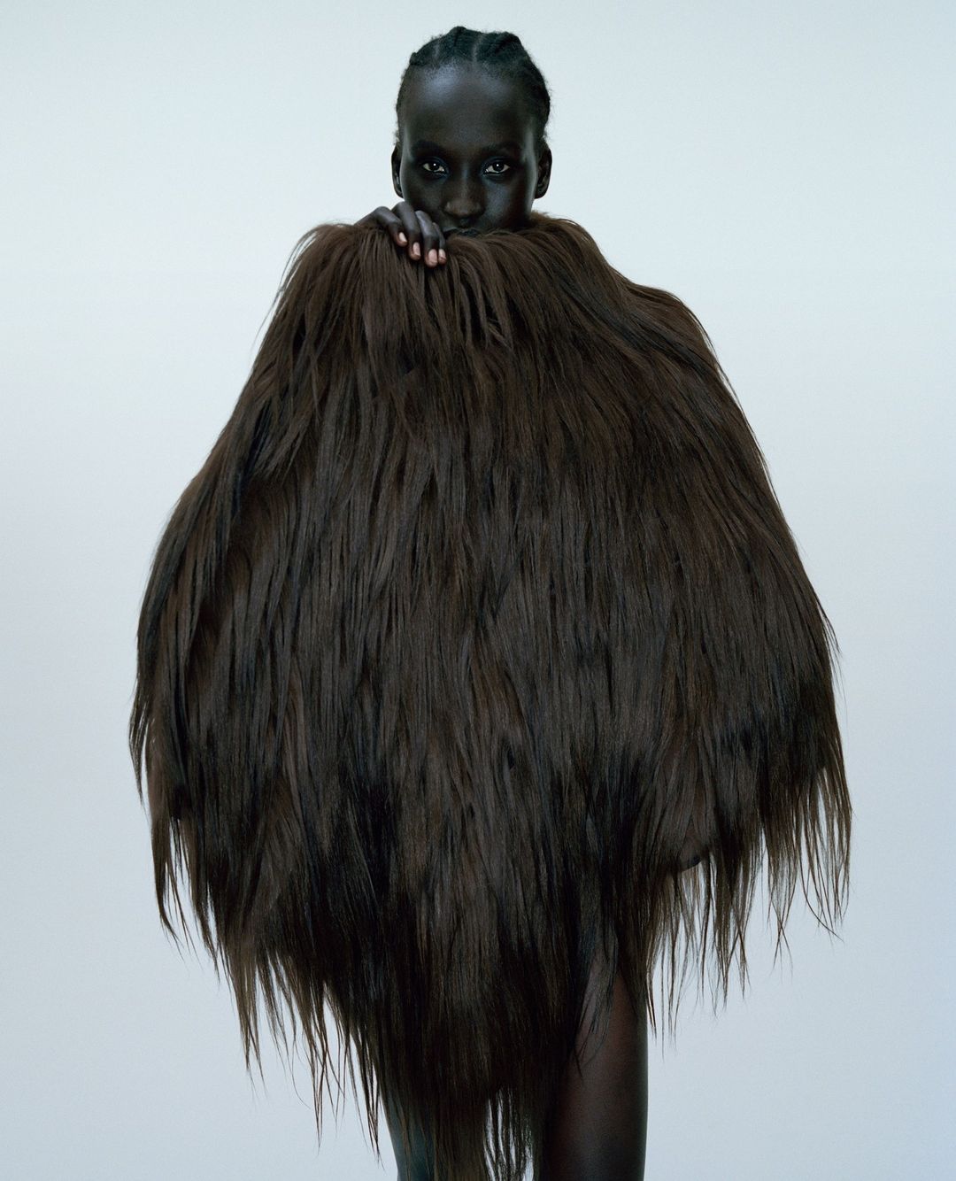 Portrait of a dark-skinned woman in a studio setting wearing a large, textured brown fur garment that covers most of her body.