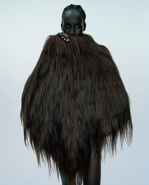 Portrait of a dark-skinned woman in a studio setting wearing a large, textured brown fur garment that covers most of her body.
