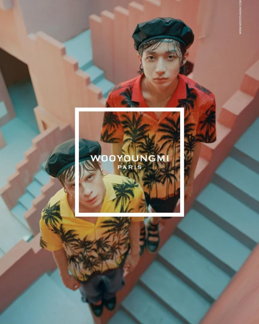 Two young adult fashion models in tropical print shirts pose on a pink staircase under soft light for a fashion editorial.