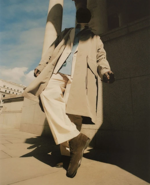 Man in a beige trench coat and tan pants strikes a pose next to a concrete column with blue sky visible in background, fashion editorial.