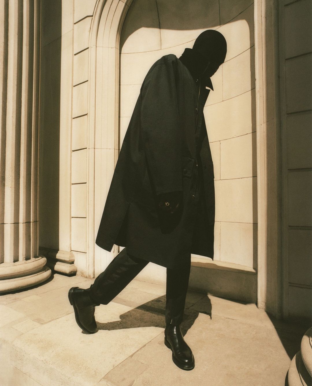 Figure in dark coat and black boots walking past classical architectural columns in London by Piczo
