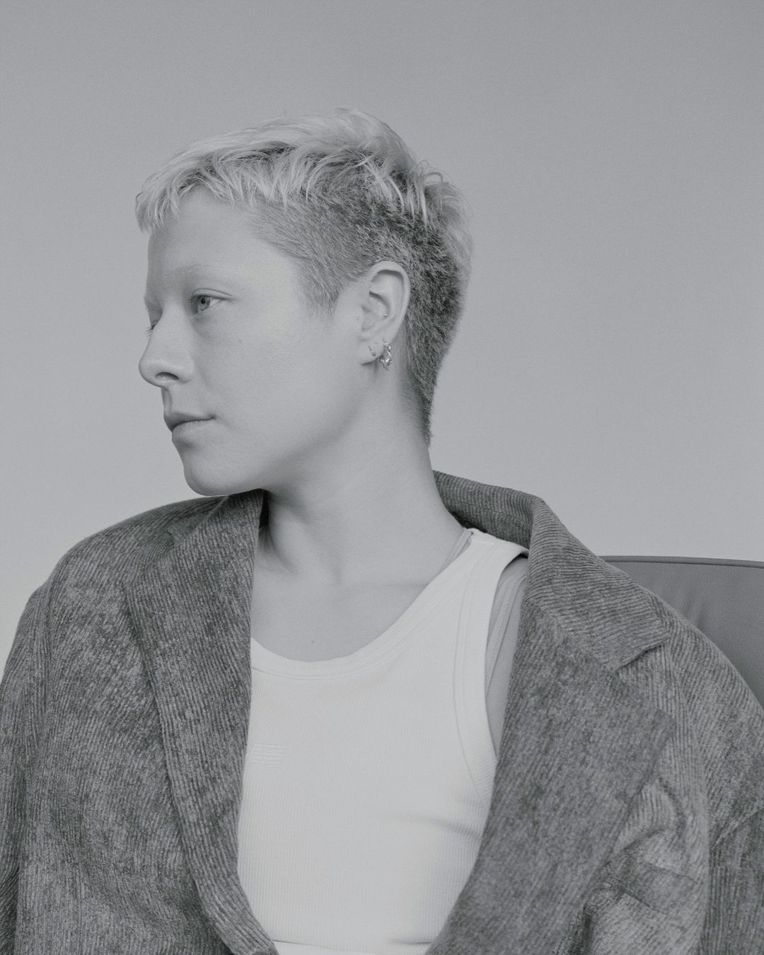 Monochrome portrait of a person with short blonde hair wearing a textured blazer and white tank top against a plain grey background