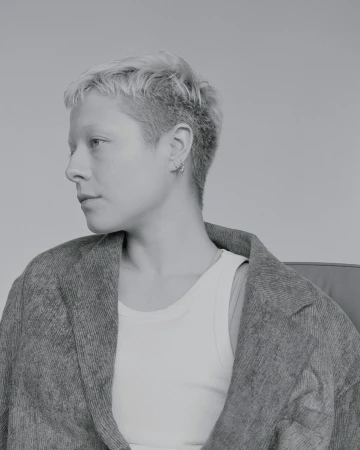 Monochrome portrait of a person with short blonde hair wearing a textured blazer and white tank top against a plain grey background