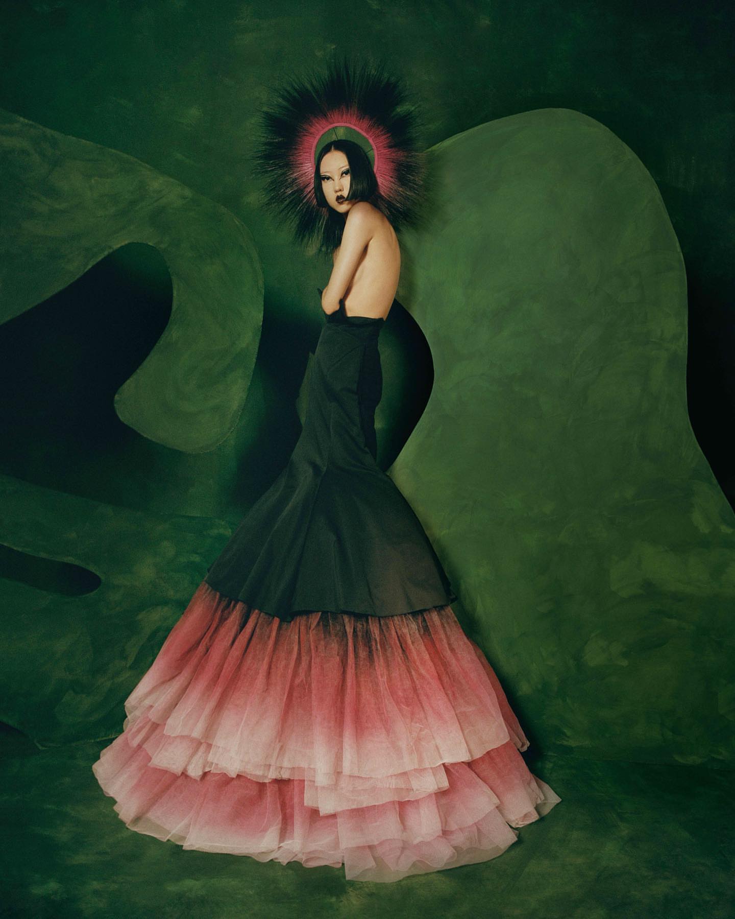 Elegant woman in a strapless black and pink ombre gown and halo posing against a textured green background in a fashion editorial.