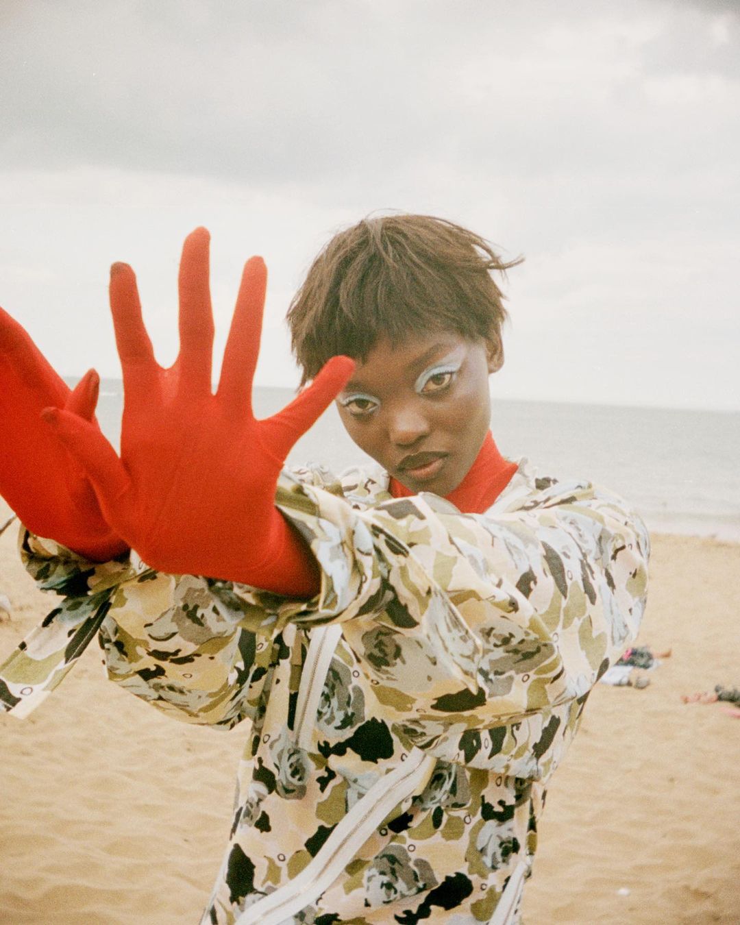 Woman with bold makeup poses on a beach, wearing a patterned jacket and red gloves, with a cloudy sky and sandy beach backdrop.