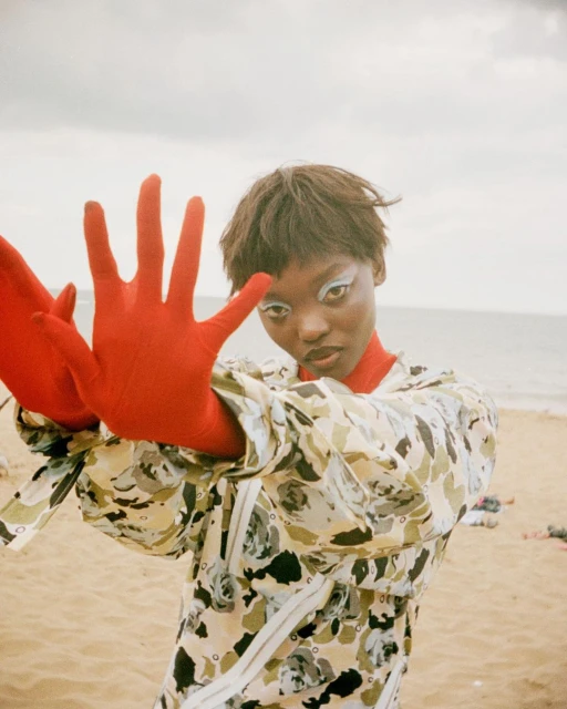 Woman with bold makeup poses on a beach, wearing a patterned jacket and red gloves, with a cloudy sky and sandy beach backdrop.