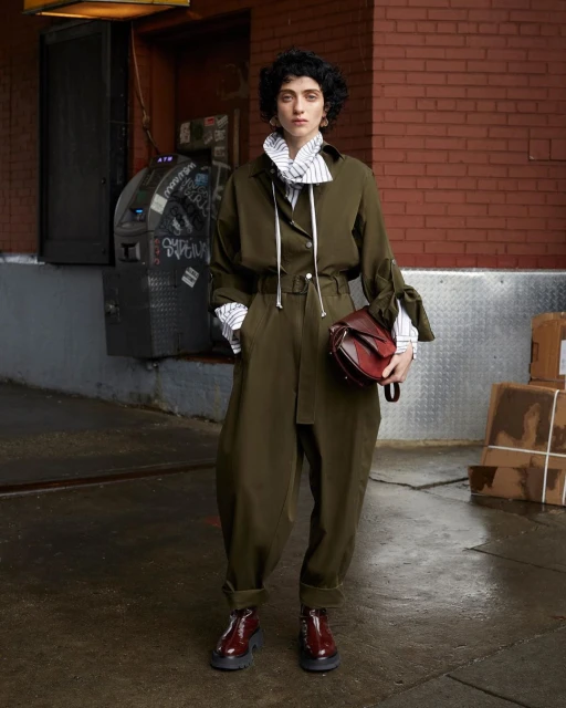 A woman poses in an olive green jumpsuit with a red handbag, complemented by burgundy shoes, against an urban brick wall backdrop.