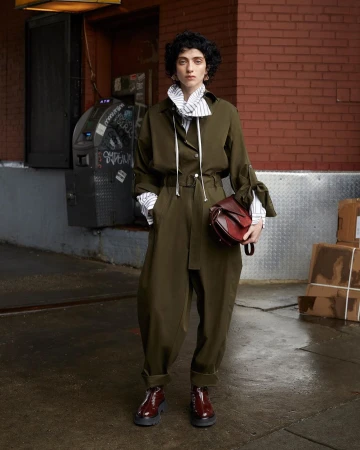 A woman poses in an olive green jumpsuit with a red handbag, complemented by burgundy shoes, against an urban brick wall backdrop.