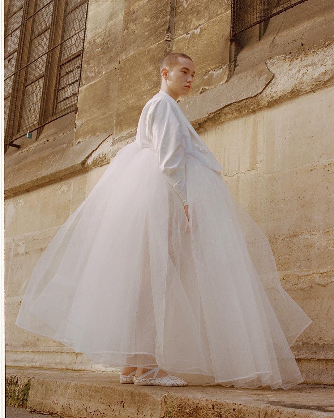 Woman in white blouse and tutu standing outdoors against a stone building, wearing ballet shoes, in editorial fashion image.