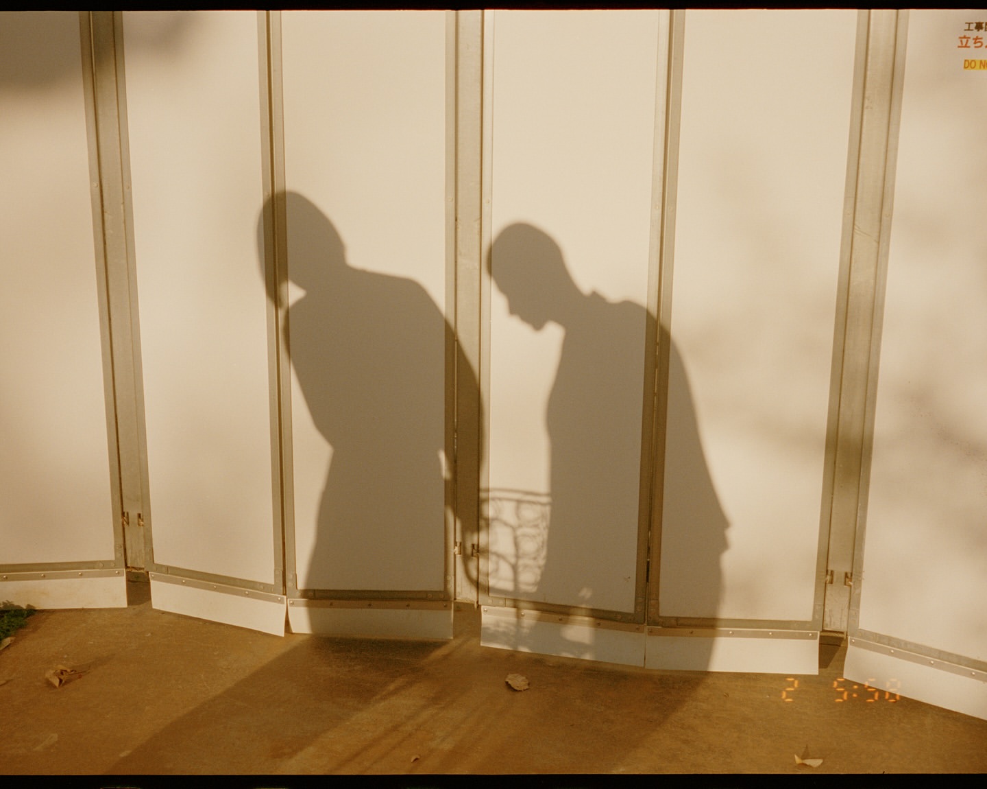 Monochrome image featuring the shadows of two people cast against a white wall with segmented panels in an indoor setting.