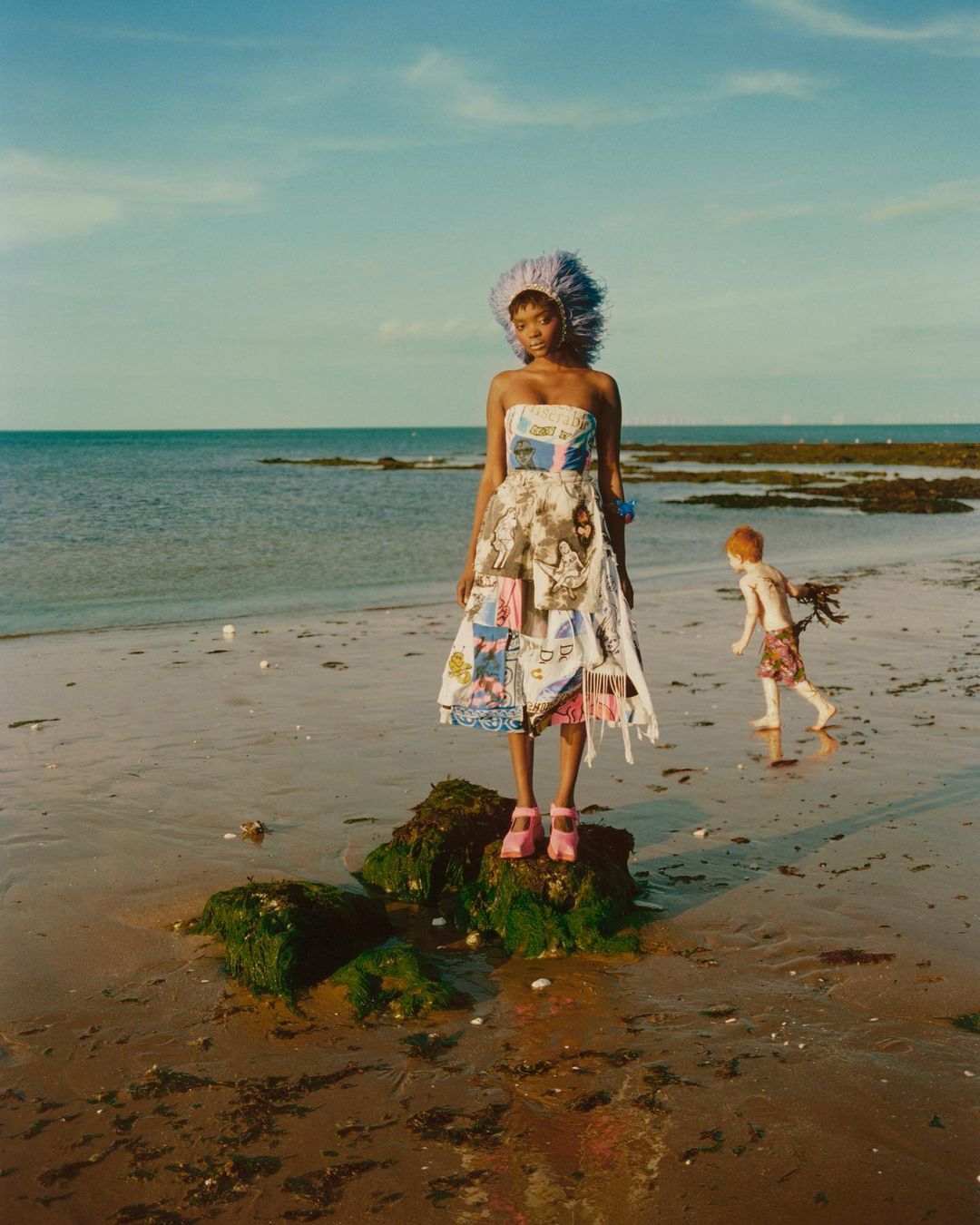A person in a patchwork dress and feathered headpiece standing on rocks at the beach, next to the ocean. A child runs in the background.