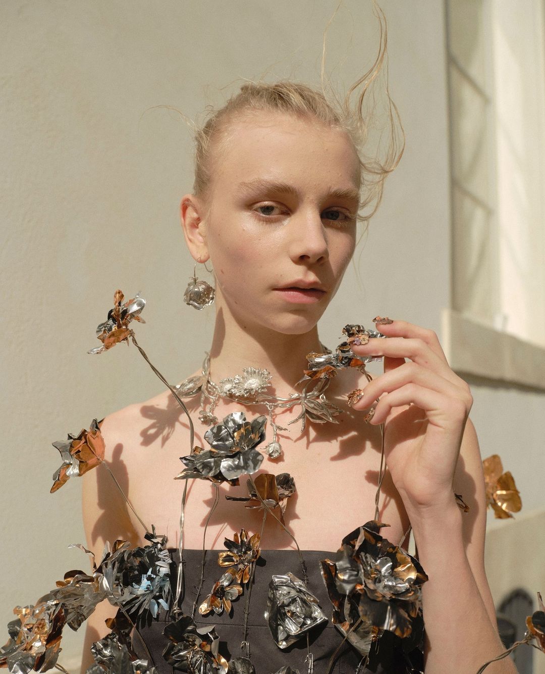 Portrait of a woman wearing a floral dress and jewelry made of flowers, posing in front of a neutral wall in natural light.