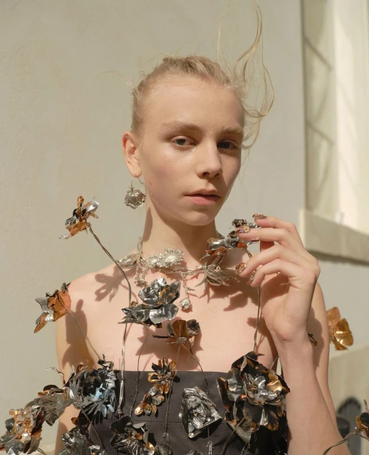 Portrait of a woman wearing a floral dress and jewelry made of flowers, posing in front of a neutral wall in natural light.