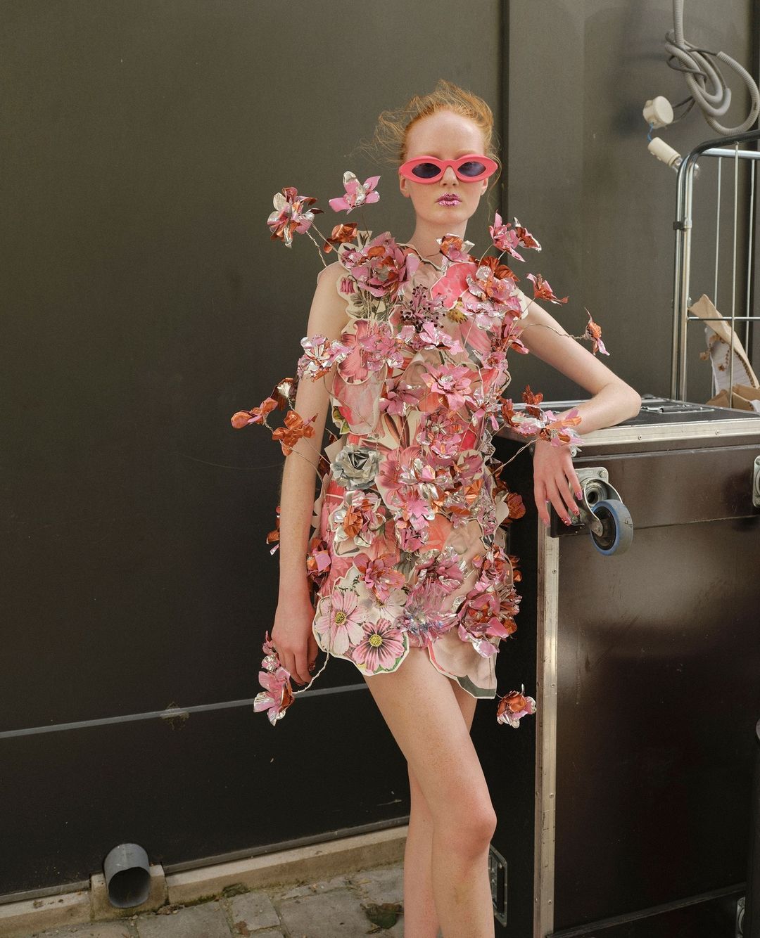 Young adult woman in pink sunglasses wearing a floral-embellished dress, posing indoors against a dark wall and equipment case.