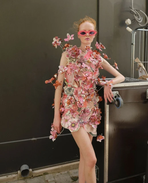 Young adult woman in pink sunglasses wearing a floral-embellished dress, posing indoors against a dark wall and equipment case.