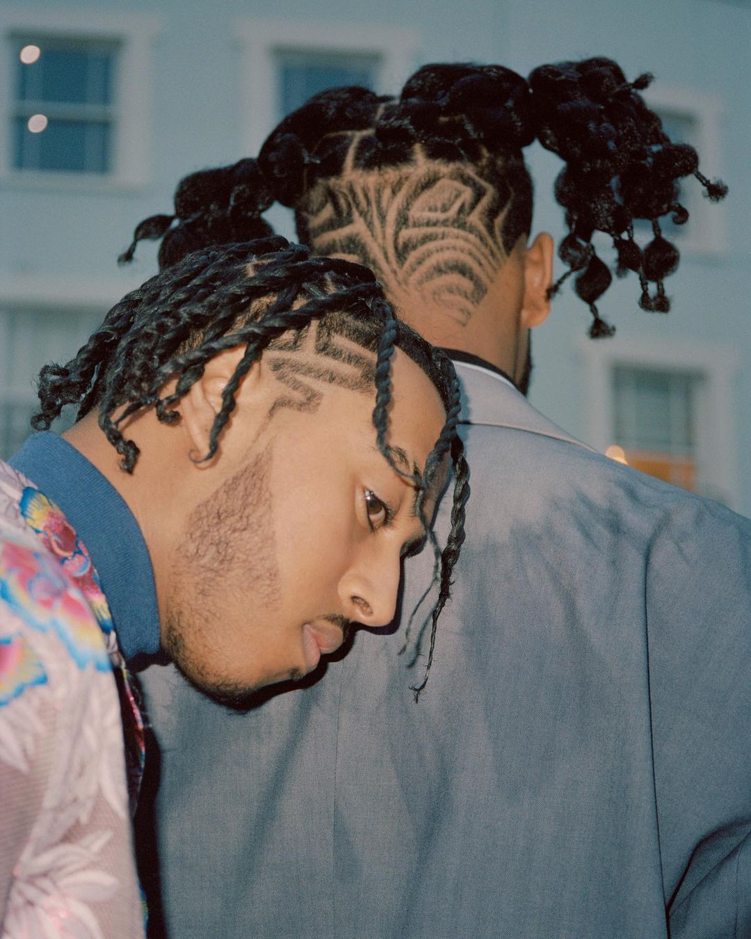 Close-up of two Black men, one with head resting on the other's shoulder, both displaying detailed and artistic haircut designs, against a building.