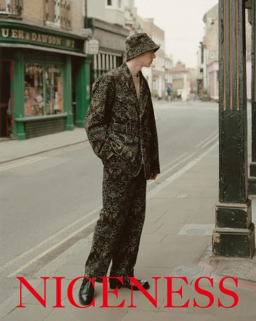 Man in patterned suit and bucket hat stands on city sidewalk next to street lamp, looking off into the distance. Fashion editorial.