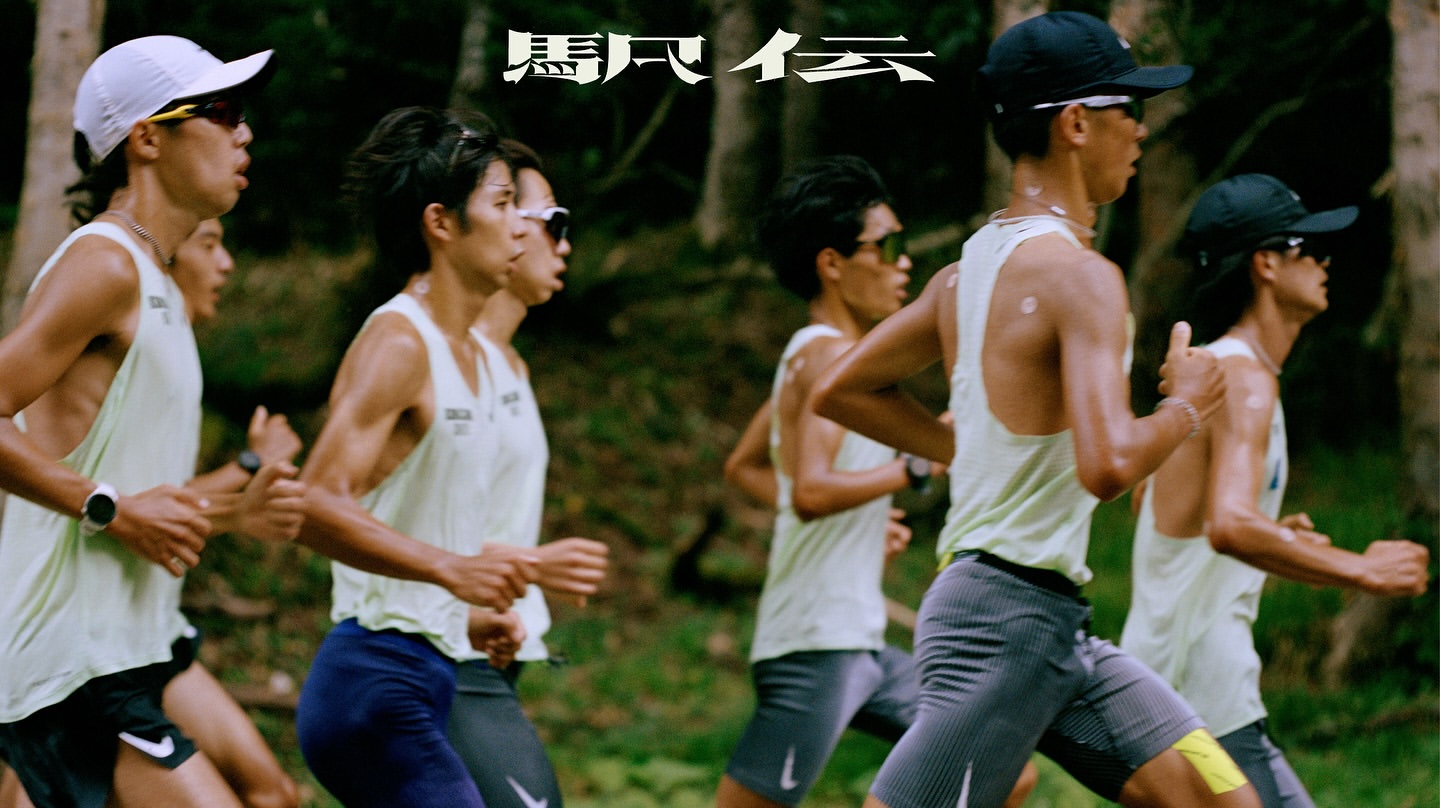 A group of male runners in athletic wear running on a path through a wooded area, shot at eye level with medium framing on a cloudy day.