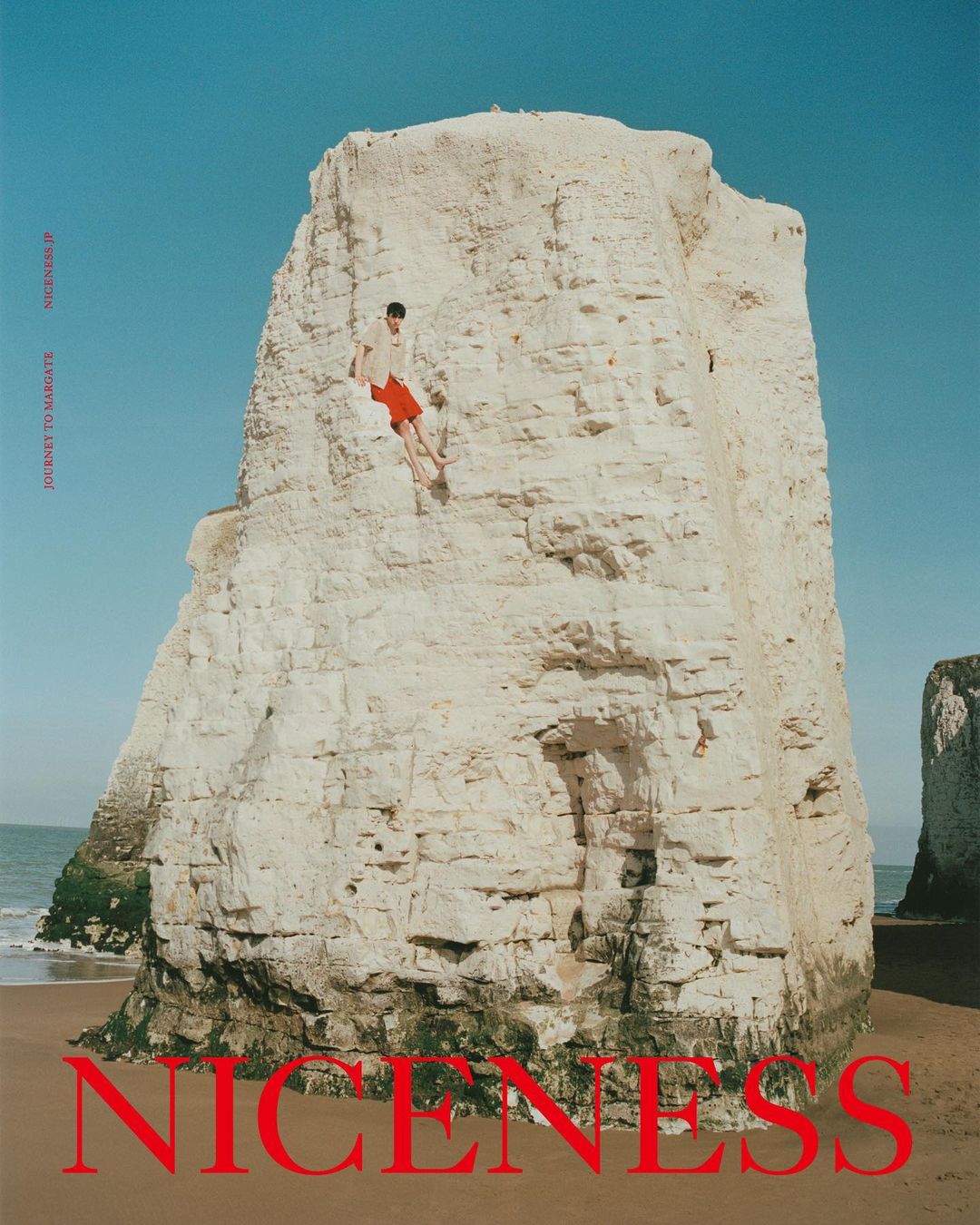 Adult male climbing a massive white rock formation on a beach under a blue sky, wearing a light top and bright red shorts.