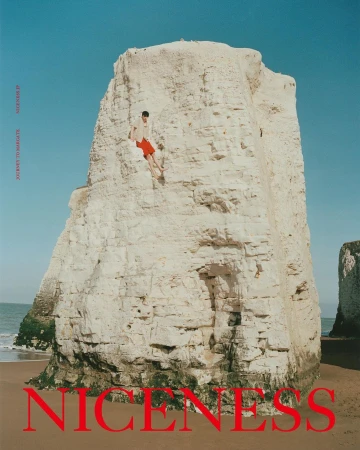Adult male climbing a massive white rock formation on a beach under a blue sky, wearing a light top and bright red shorts.