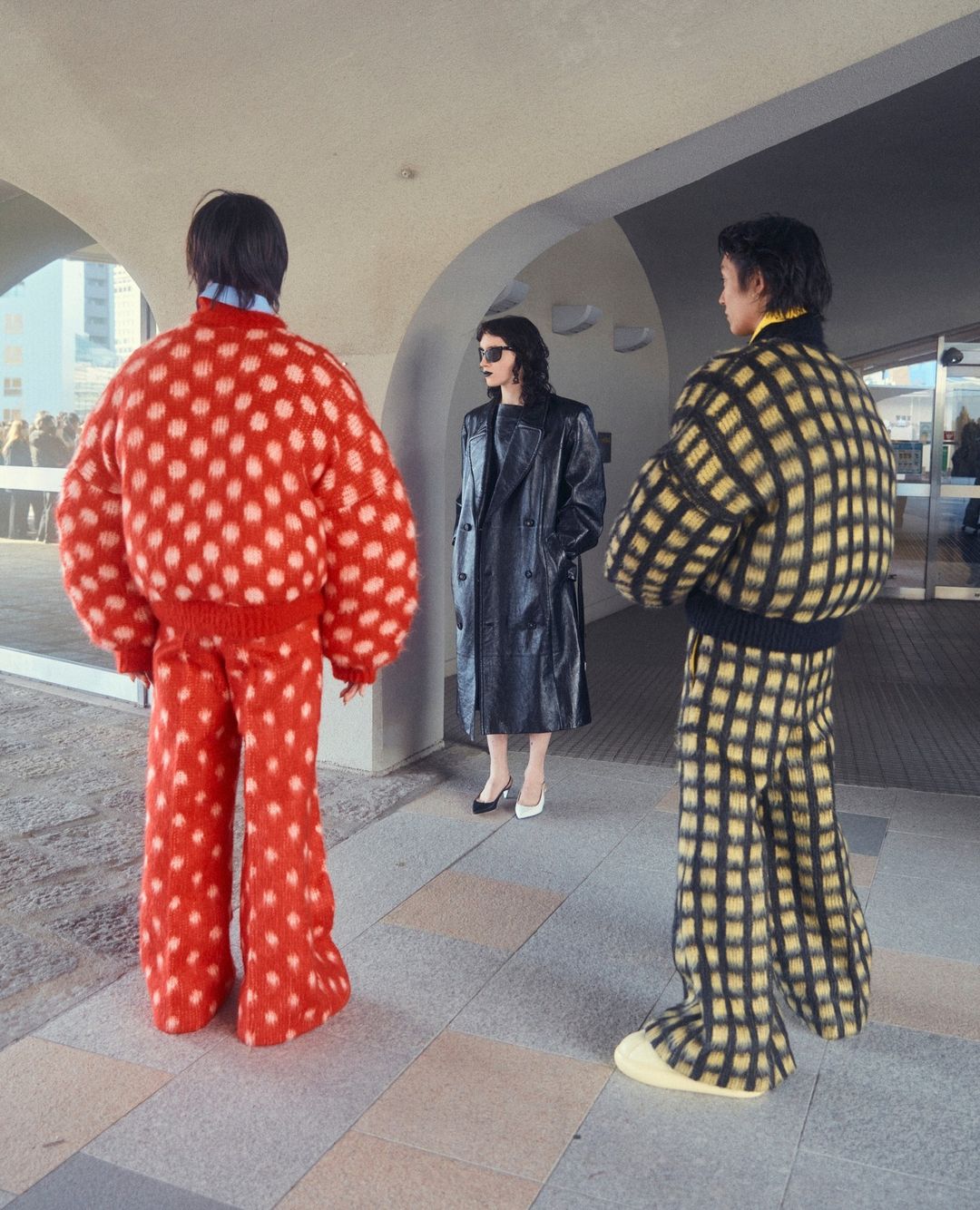 Three people in patterned outfits stand outside a modern building; one wears a red polka-dot suit, another wears a checkered suit, and another a black coat.