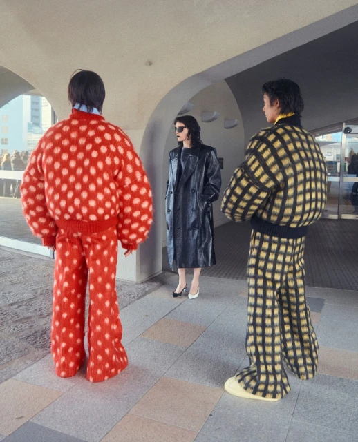 Three people in patterned outfits stand outside a modern building; one wears a red polka-dot suit, another wears a checkered suit, and another a black coat.