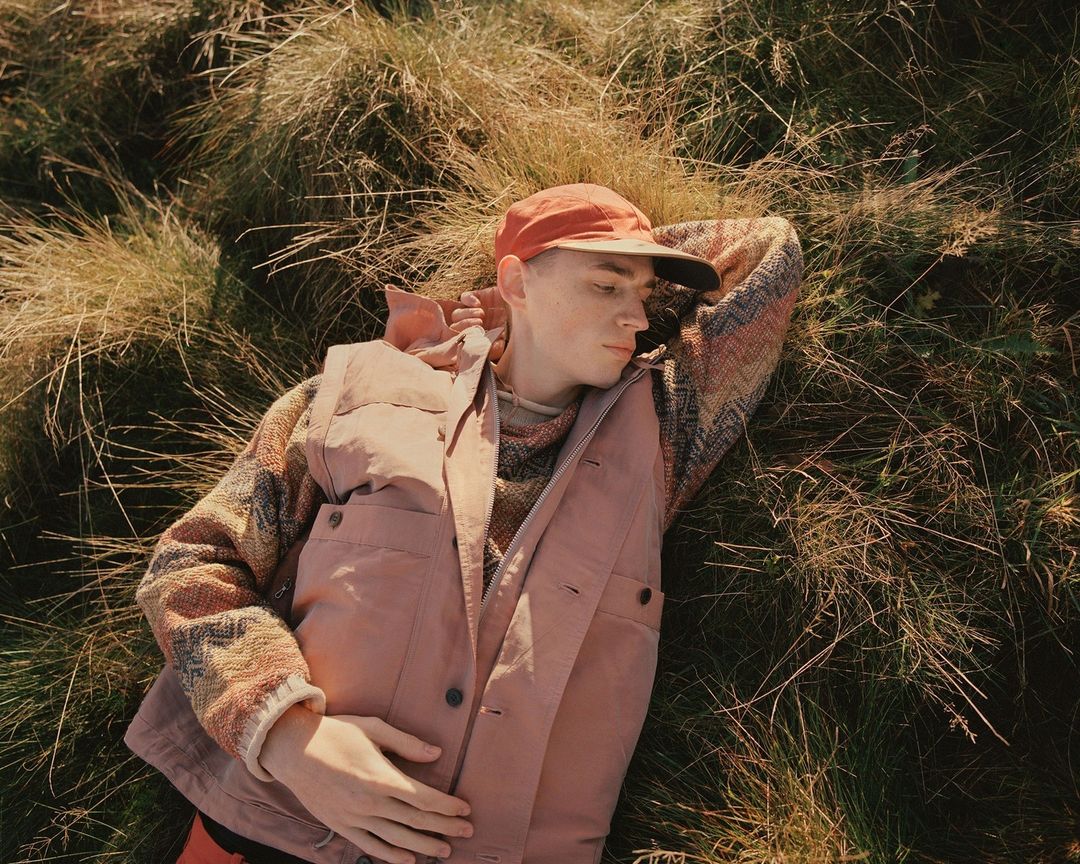 A person lies in tall grass, wearing an orange cap, patterned sweater, and pink vest, in an eye-level shot.