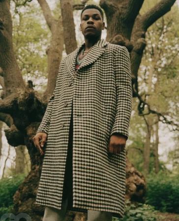 Portrait of John Boyega standing outdoors in a houndstooth overcoat before a large gnarled tree in woodland shot by Piczo