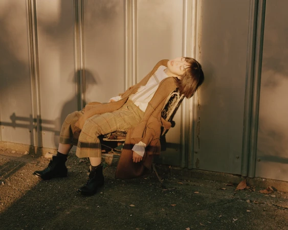 Woman in brown jacket and plaid pants lounges in chair outdoors with sunlight casting shadows against metal wall. Boots visible.