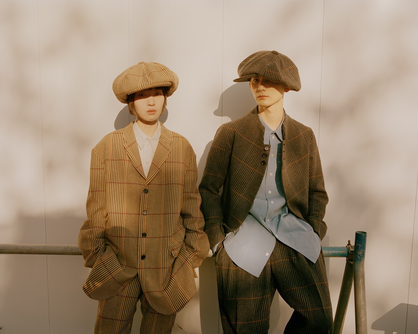 Two people wearing matching brown plaid suits and newsboy caps standing side-by-side against a white wall in soft, natural lighting.