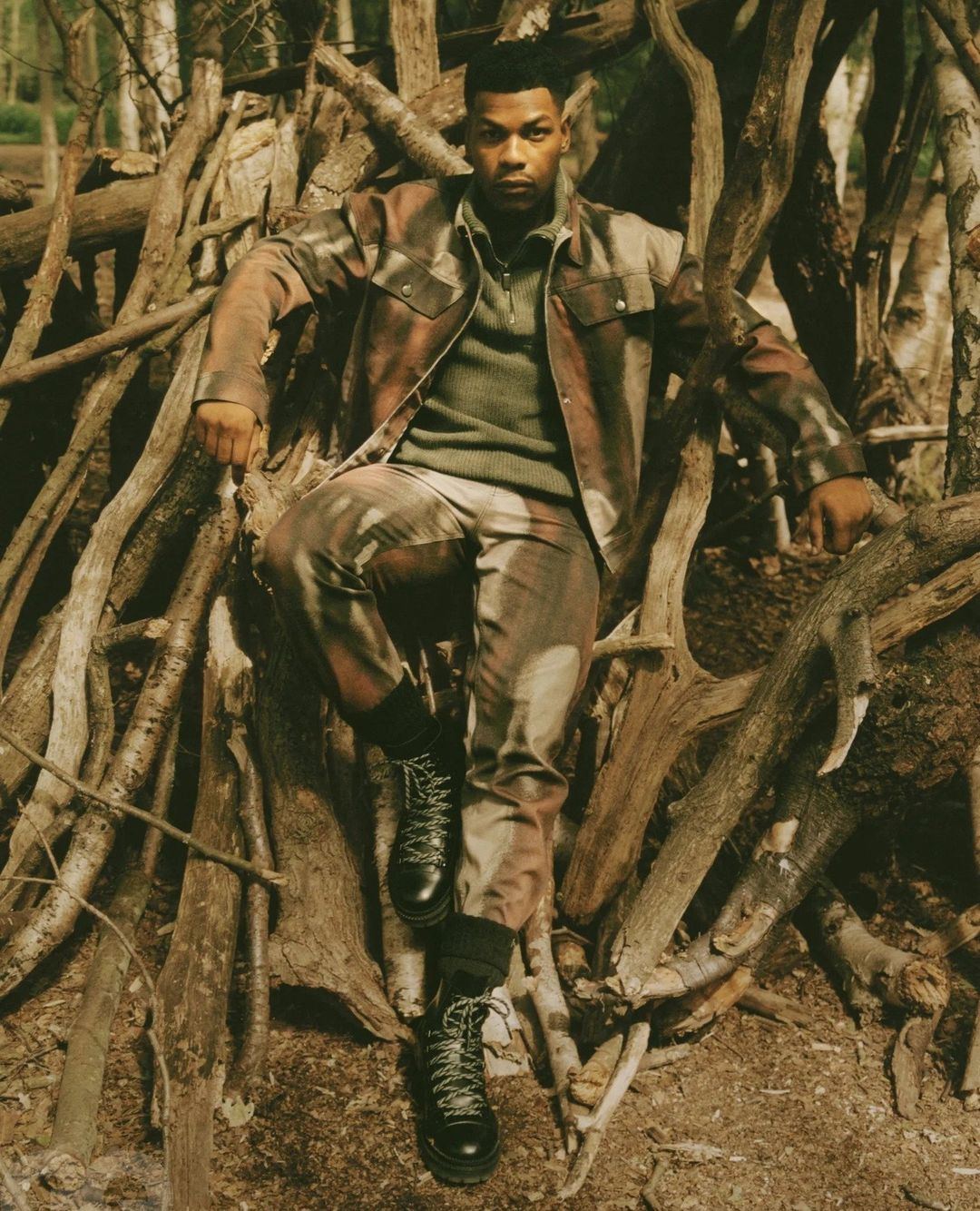 John Boyega seated amid a structure of stacked logs wearing a camouflage jacket and trousers by photographer Piczo.