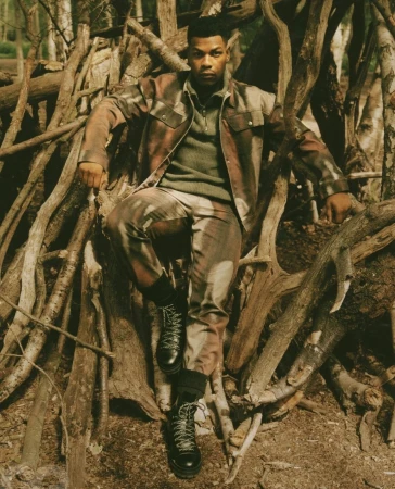 John Boyega seated amid a structure of stacked logs wearing a camouflage jacket and trousers by photographer Piczo.