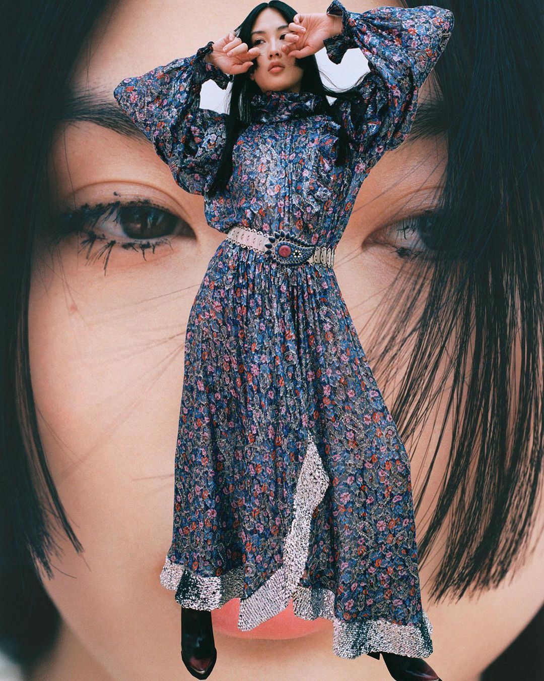 Woman poses in a floral dress with a large face in the background. She is wearing a floral maxi dress with a wide belt.